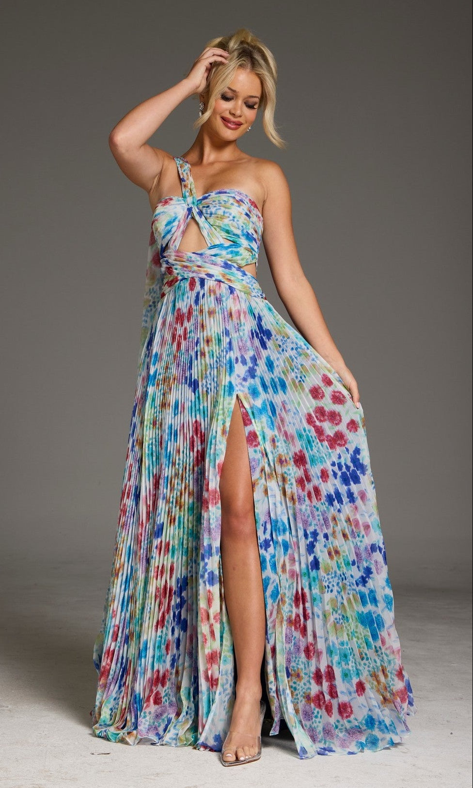 A woman in the Jovani 38689 long prom dress in print, featuring a colorful pleated A-line shape, one-shoulder design, front cutout, and thigh-high slit, stands against a plain background holding part of the skirt.